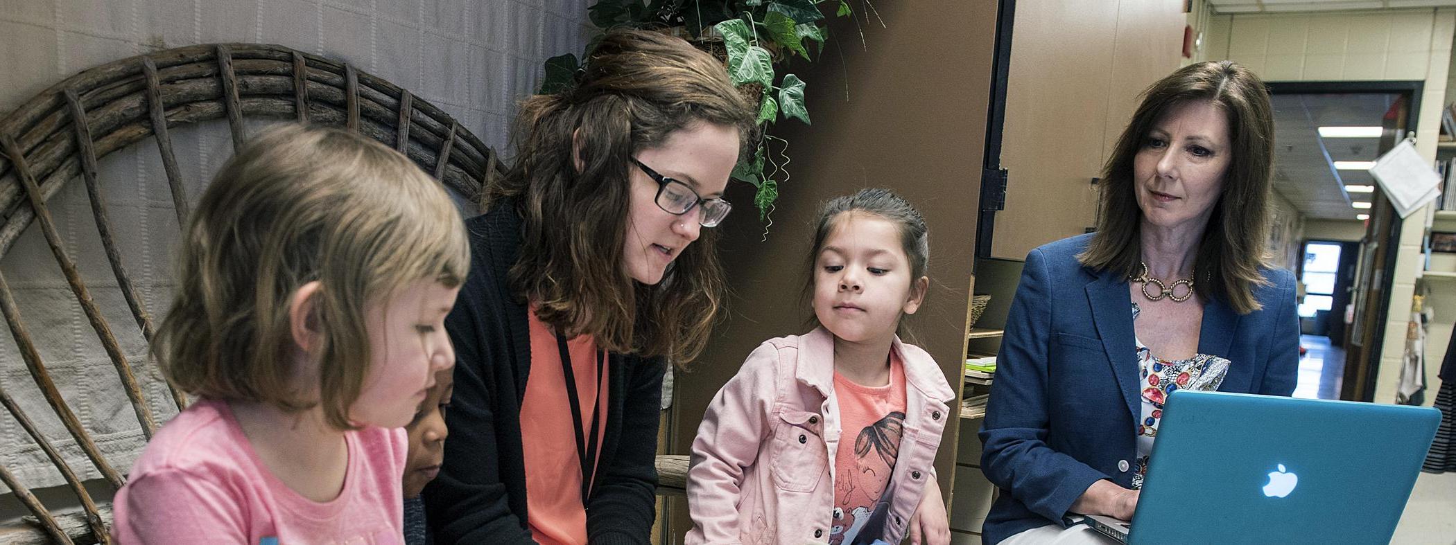 A female student teacher reads to children while faculty looks on